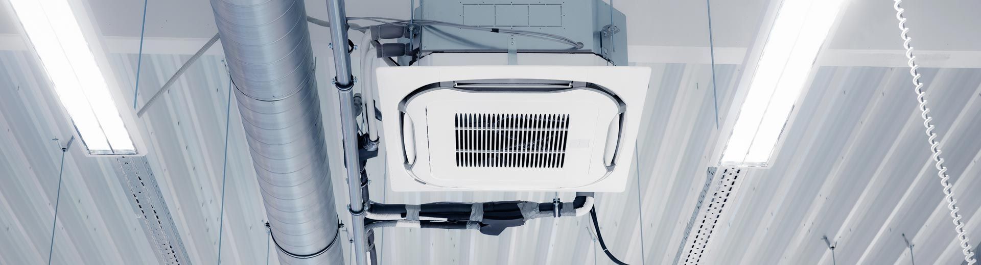 Unité de climatisation blanche fixée sur un plafond métallique avec des tubes fluorescents et des tuyaux.