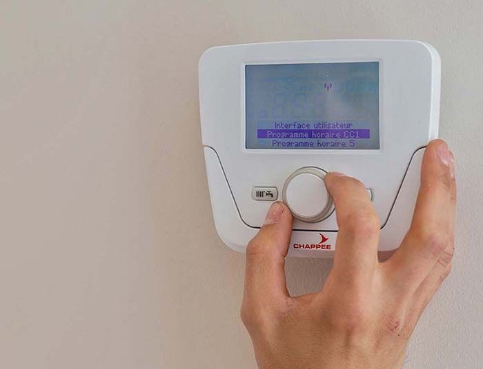 Main tournant un cadran sur un thermostat blanc monté sur un mur beige, affichant un menu sur l'écran.