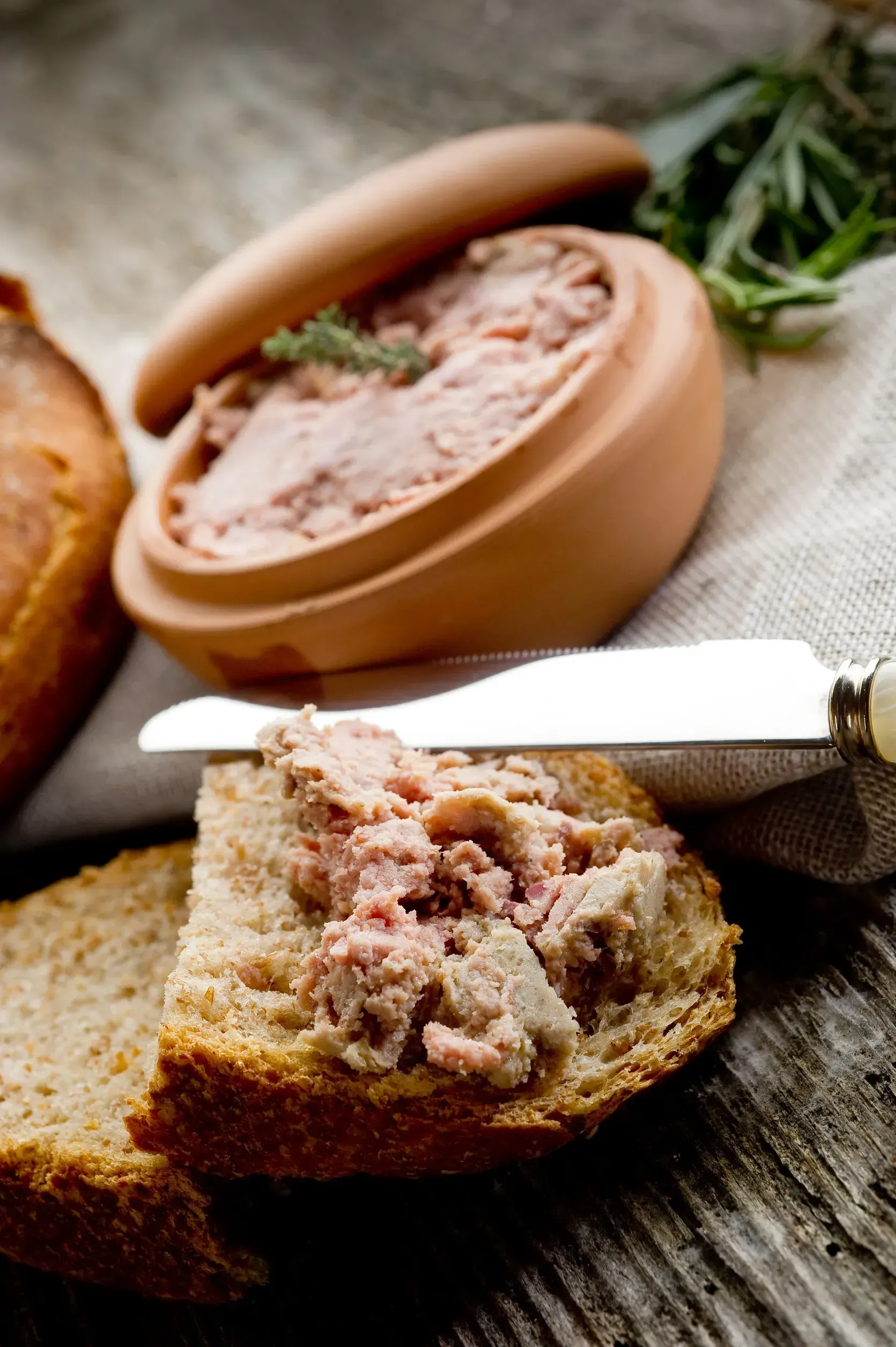 Paté untado con cuchillo sobre pan tostado, servido junto a un cazo de cerámica y una hogaza de pan.