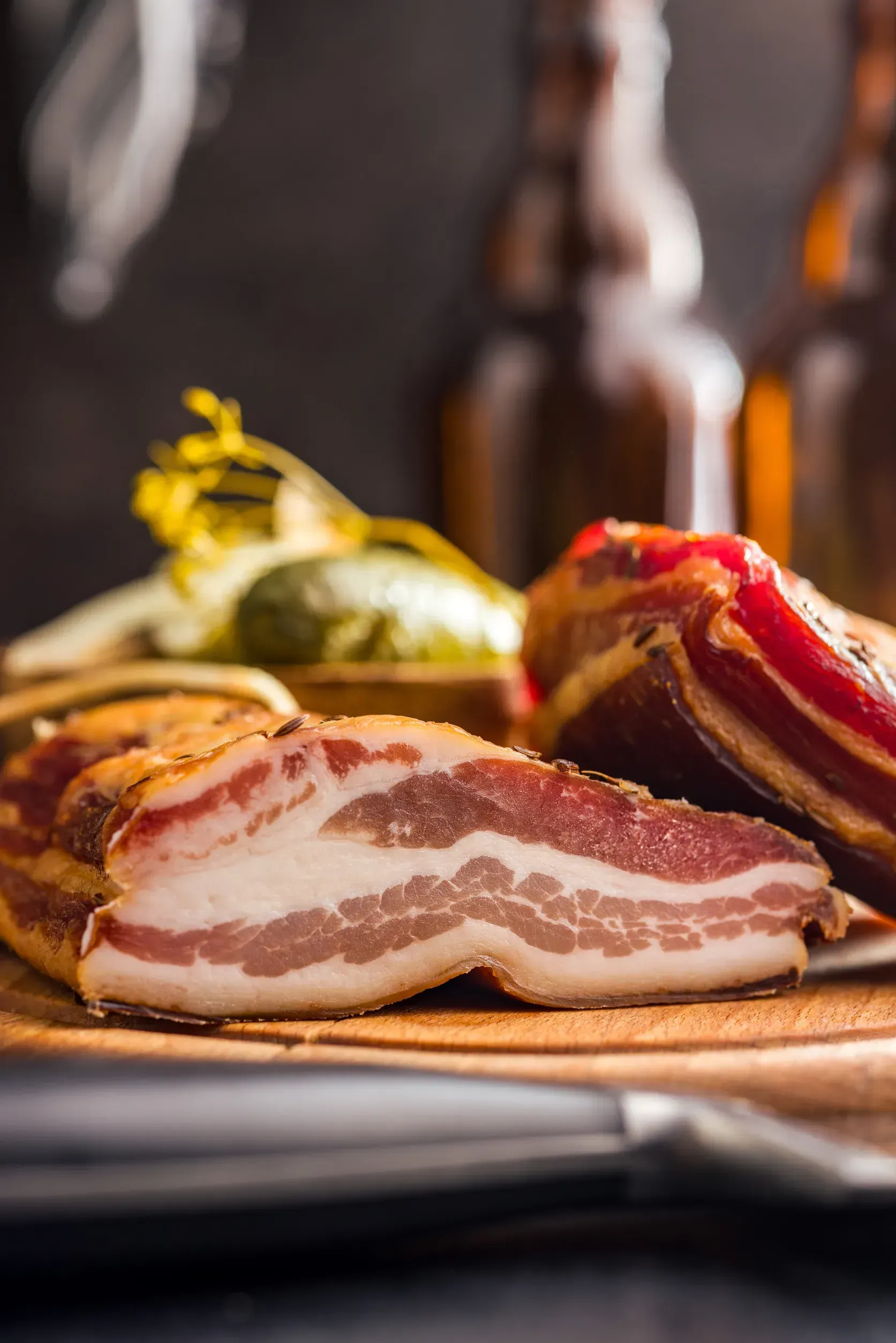 Primer plano de carne curada sobre una tabla de madera, con encurtidos y botellas marrones borrosas en el fondo.