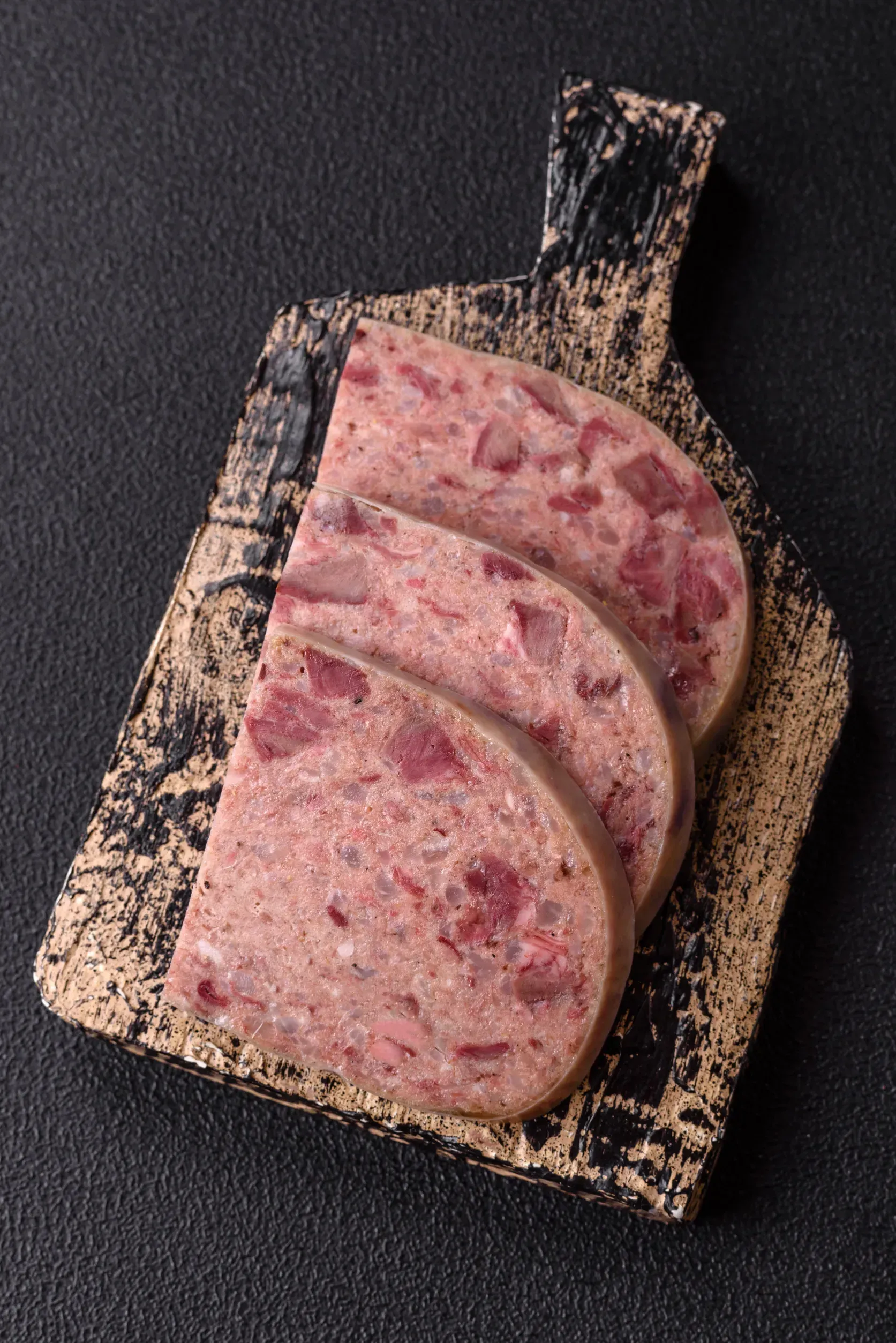 Rebanadas de salchicha de hígado con trozos de carne visibles sobre una tabla de cortar de madera texturizada, fondo negro.