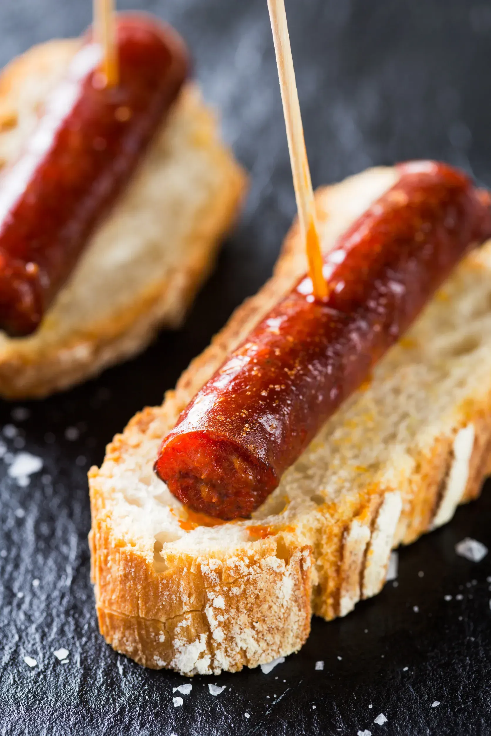 Chorizo sobre pan tostado, ensartado con un palillo sobre una superficie oscura.