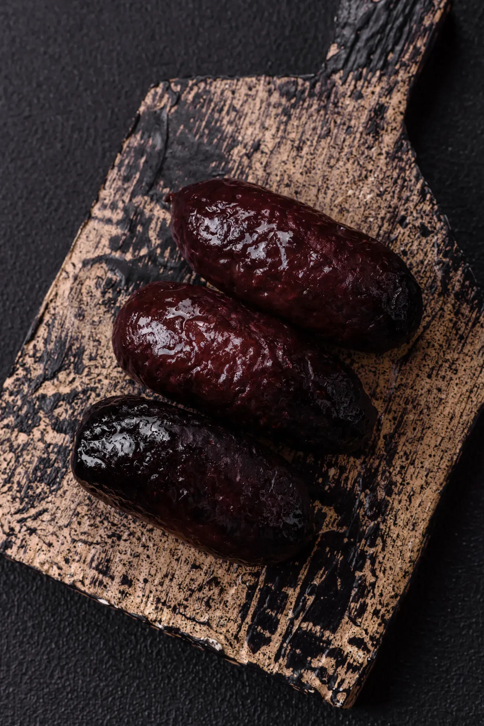 Tres pasteles glaseados de color marrón oscuro sobre una tabla de cortar texturizada de color marrón oscuro y tostado contra un fondo negro.