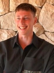 Mustaan ​​napitettavaan paitaan pukeutunut henkilö hymyilee vaalean kivimuurin edessä.