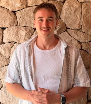 Viiksikäs henkilö hymyilee ruskean kivimuurin edessä yllään valkoinen t-paita ja napittamaton kuviollinen paita.