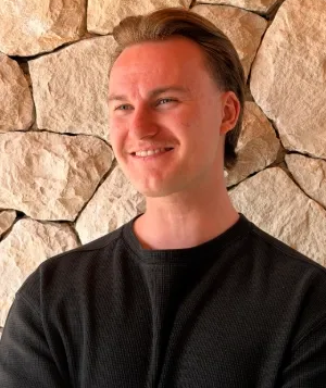 Hymyilevä, vaaleanruskeatukkainen henkilö mustassa villapaidassa vaalean kivimuurin edessä.