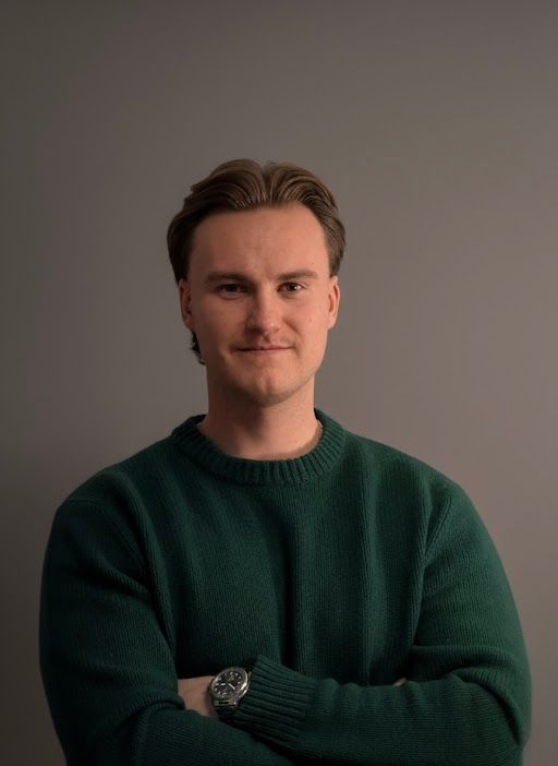Hymyilevä, vaaleanruskeatukkainen henkilö mustassa villapaidassa vaalean kivimuurin edessä.