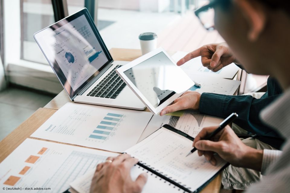 Zwei Personen arbeiten an einem Tisch mit Laptop, Tablet und ausgedruckten Diagrammen zusammen; eine Person zeigt auf etwas