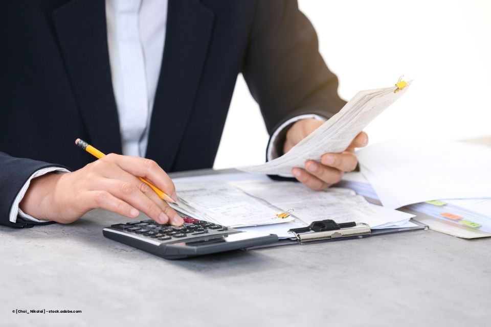 Eine Person im Businessanzug benutzt einen Taschenrechner, während sie Finanzdokumente auf einem Klemmbrett durchsieht.