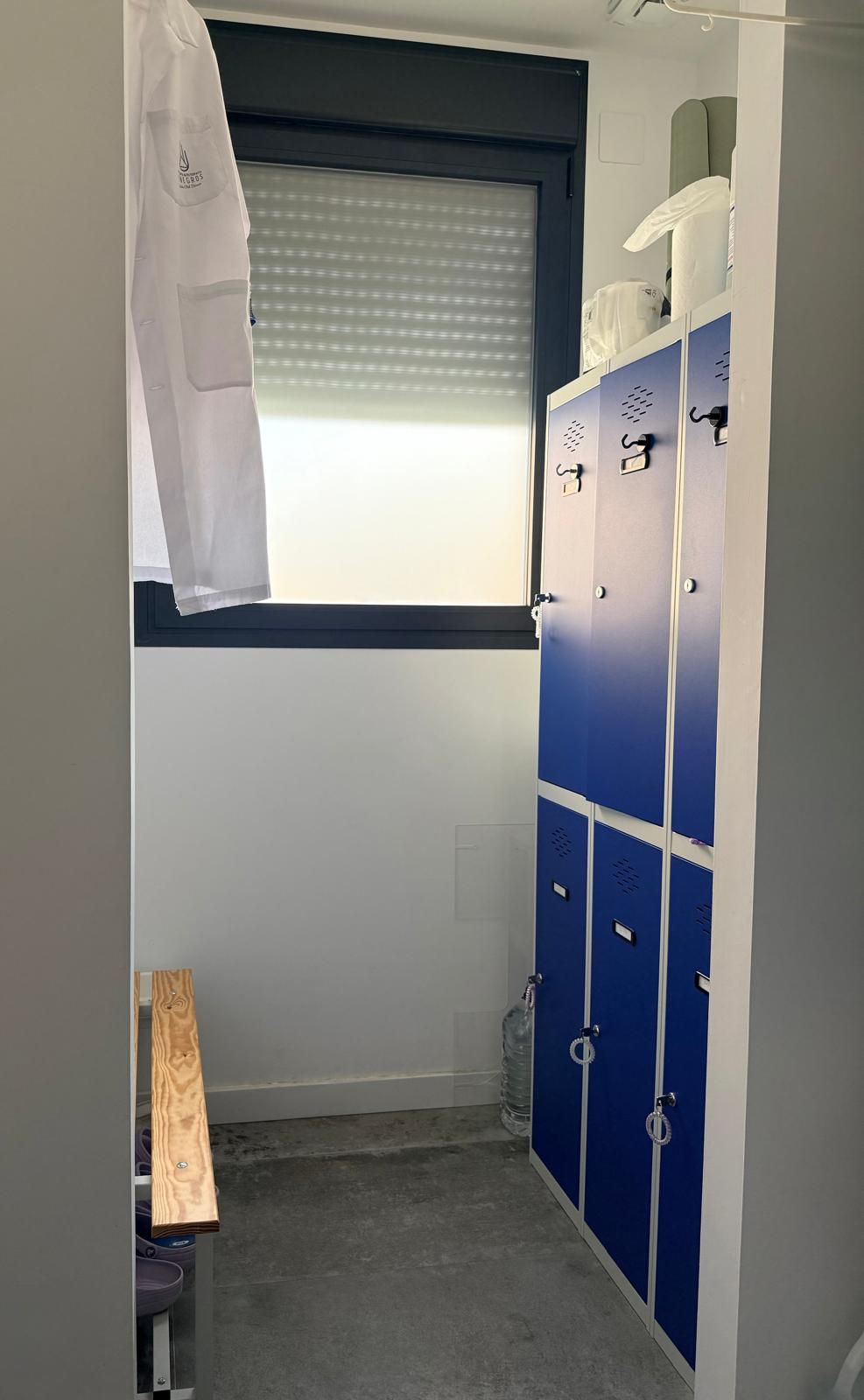 Vestuario estrecho con taquillas azules, un banco y una ventana con una persiana parcialmente cerrada.