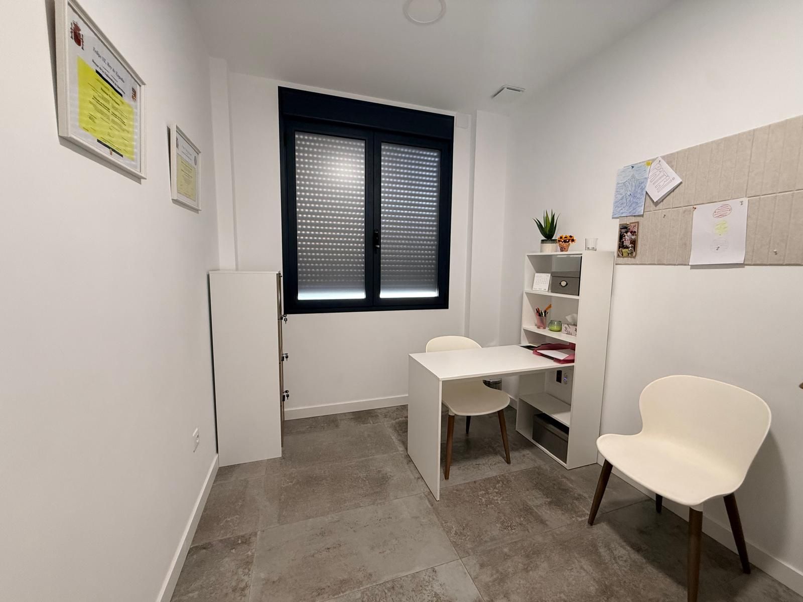 Pequeña oficina con escritorio blanco, silla, estantes y una ventana negra en una habitación minimalista.