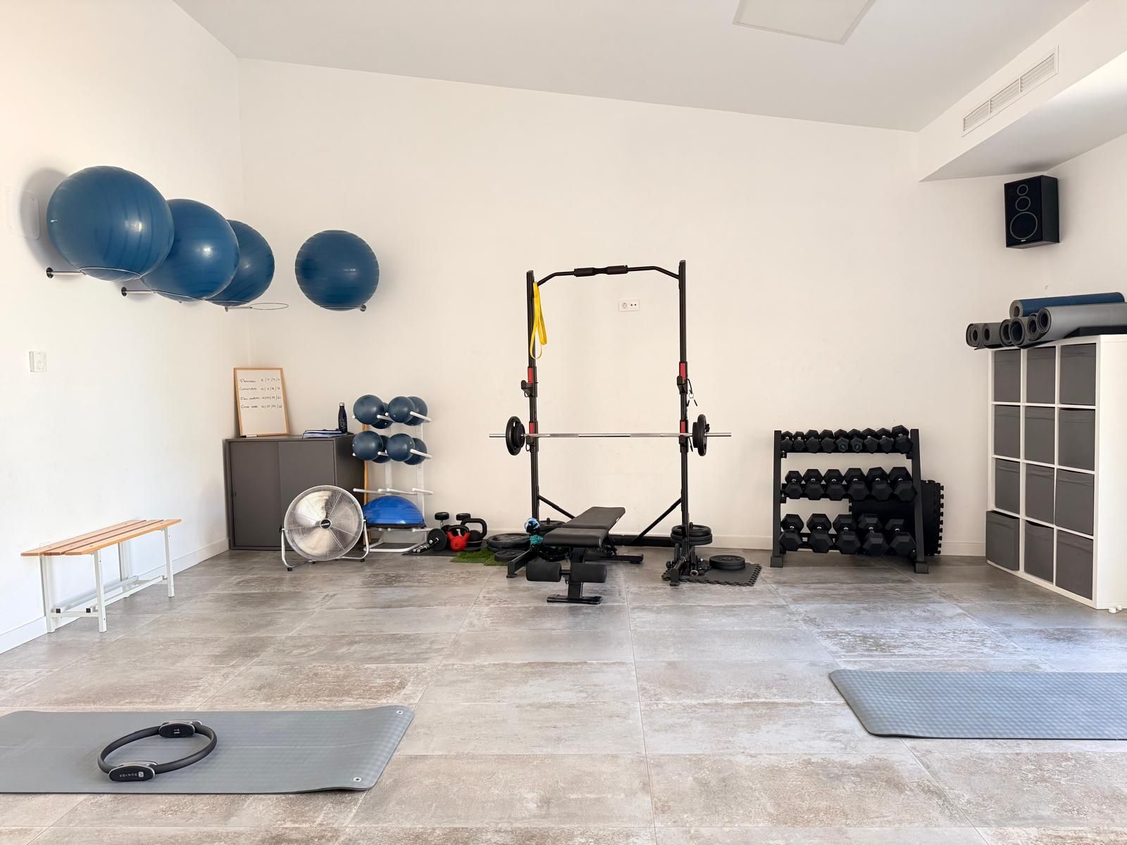 Gimnasio en casa con balones de ejercicio, mancuernas, banco, colchoneta y almacenamiento montado en la pared en una habitación luminosa.