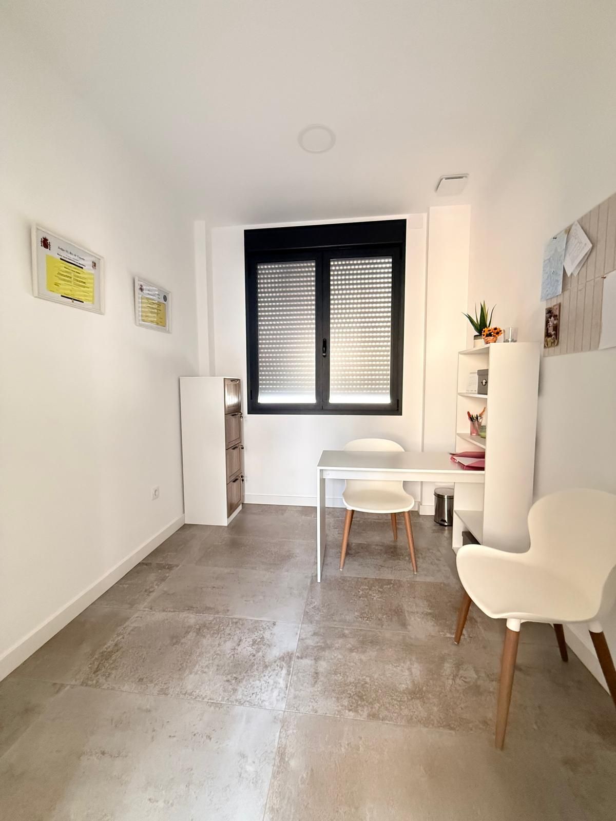 Habitación luminosa y minimalista con paredes blancas, escritorio, silla, ventana y suelo de hormigón.