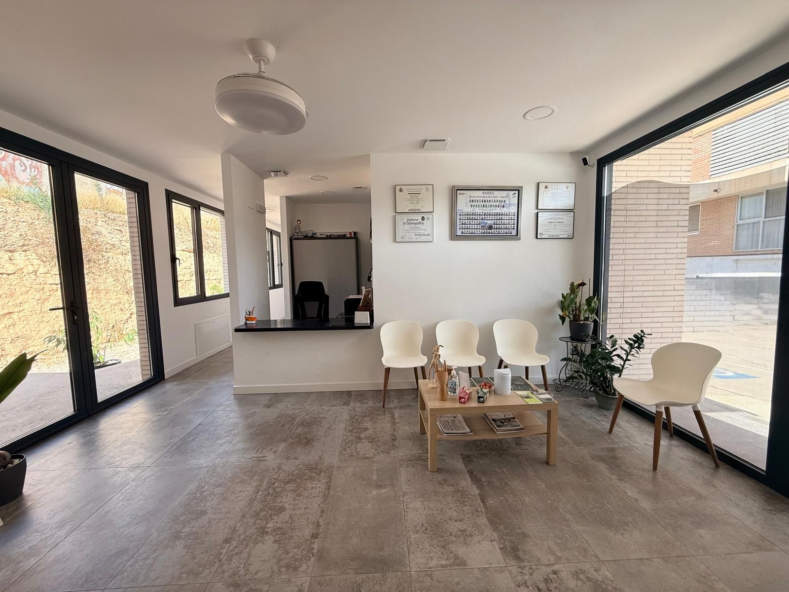 Moderna sala de espera con grandes ventanales, sillas blancas, una mesa de centro y un mostrador de recepción.