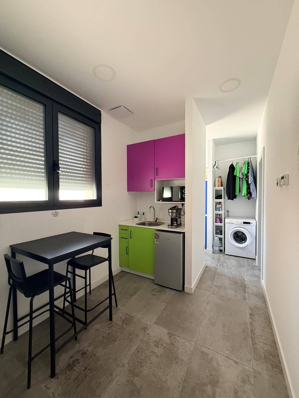 Cocina pequeña con armarios rosas y verdes, una mesa de comedor y un pasillo que conduce a la zona de lavandería.