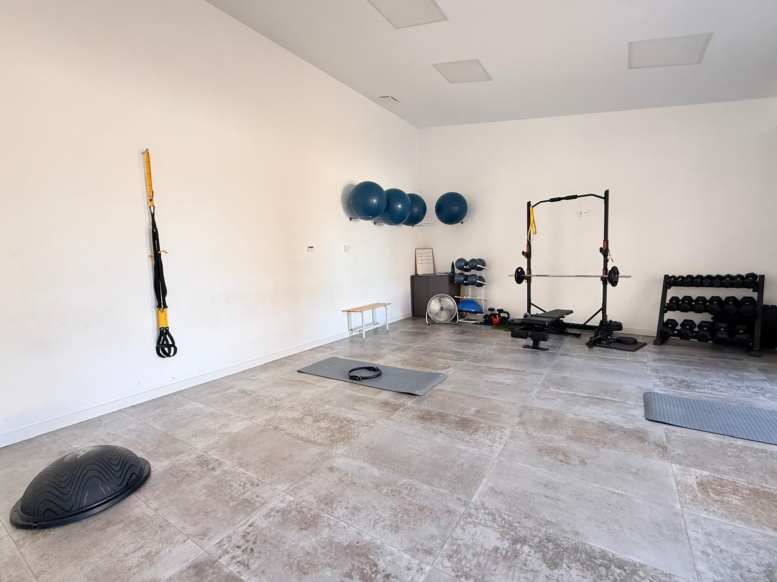 Gimnasio vacío con suelo de goma, balones de ejercicio, mancuernas, banco y una jaula de sentadillas contra paredes blancas.