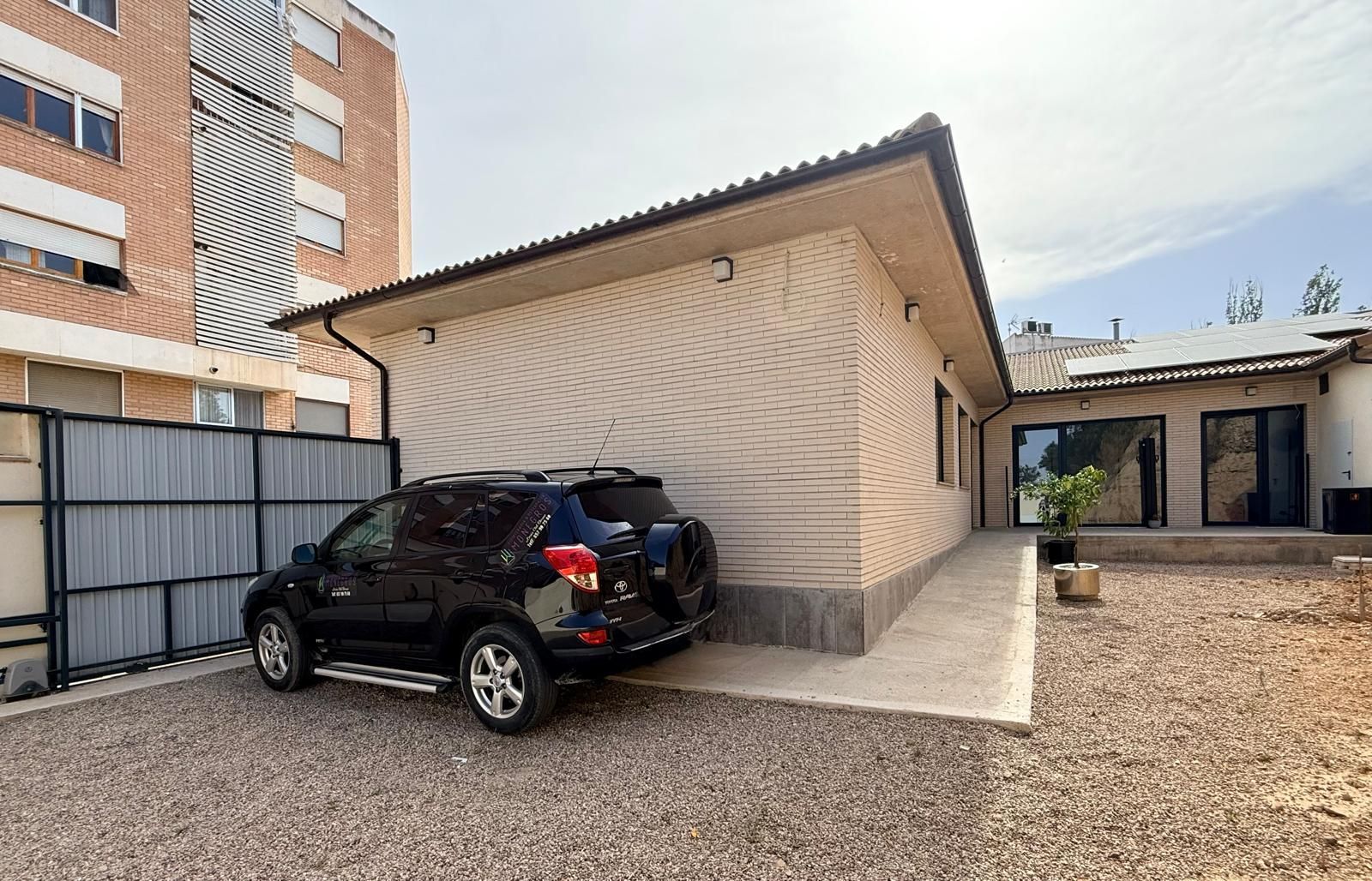 Un SUV negro estacionado junto a una casa beige en un camino de grava bajo un cielo parcialmente nublado.