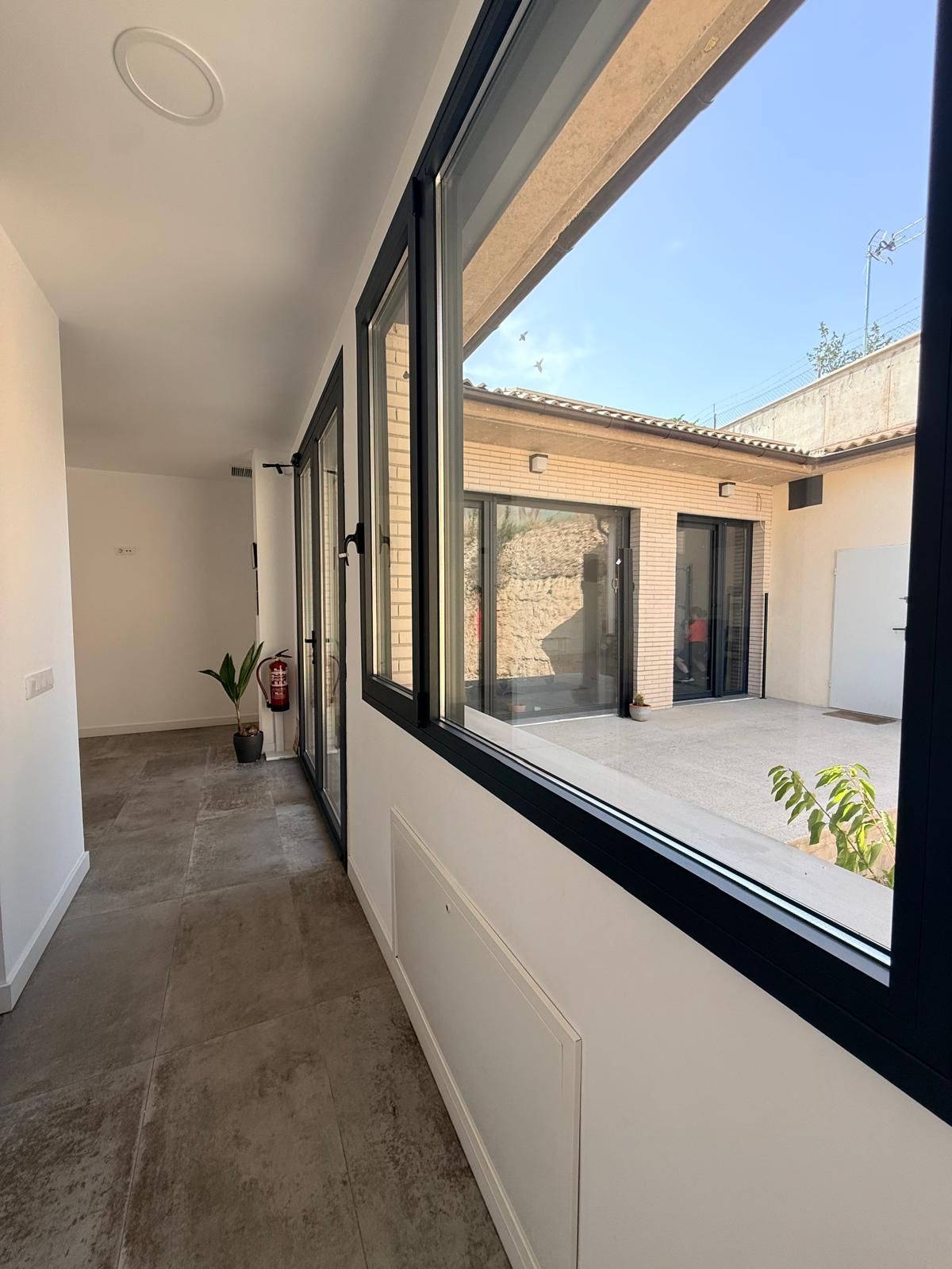 Pasillo luminoso con grandes ventanales que dan a un patio soleado y a un edificio de ladrillo.
