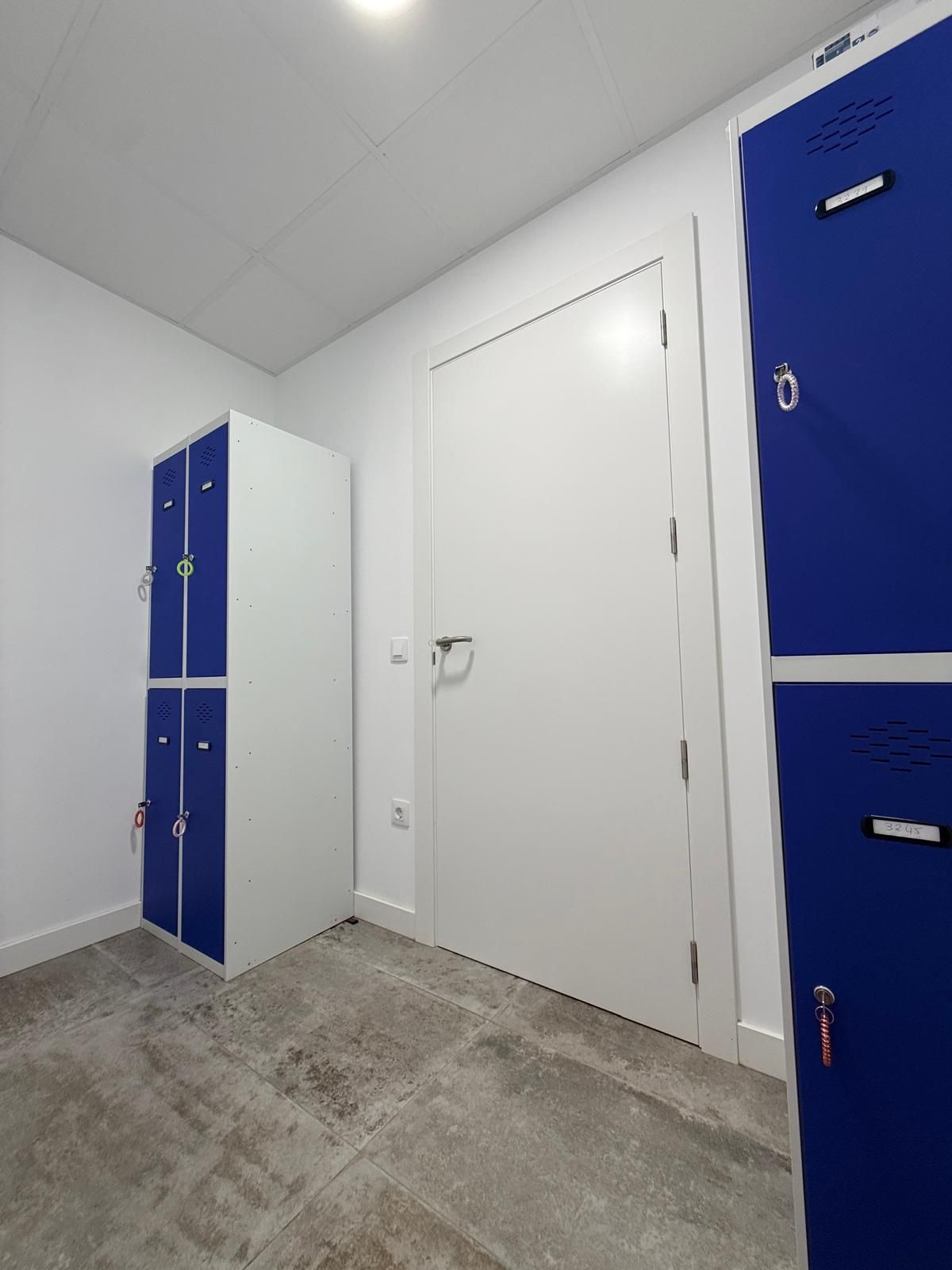 Trastero blanco con taquillas azules, una puerta blanca cerrada y suelo de hormigón gris.