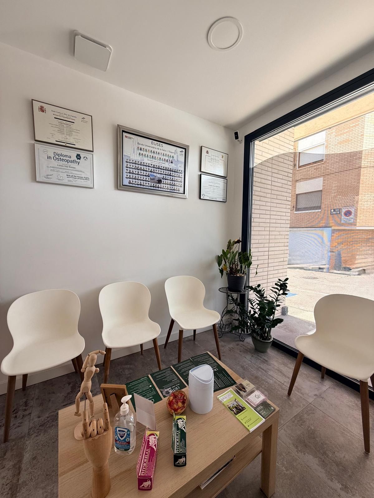 Sala de espera moderna con sillas blancas, una mesa de centro, obras de arte en la pared y una gran ventana con vistas a edificios de ladrillo.