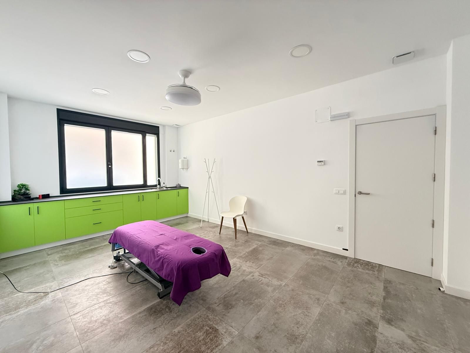 Sala de terapia luminosa con armarios verdes, camilla de masaje morada, sillas blancas y una pared con ventana.