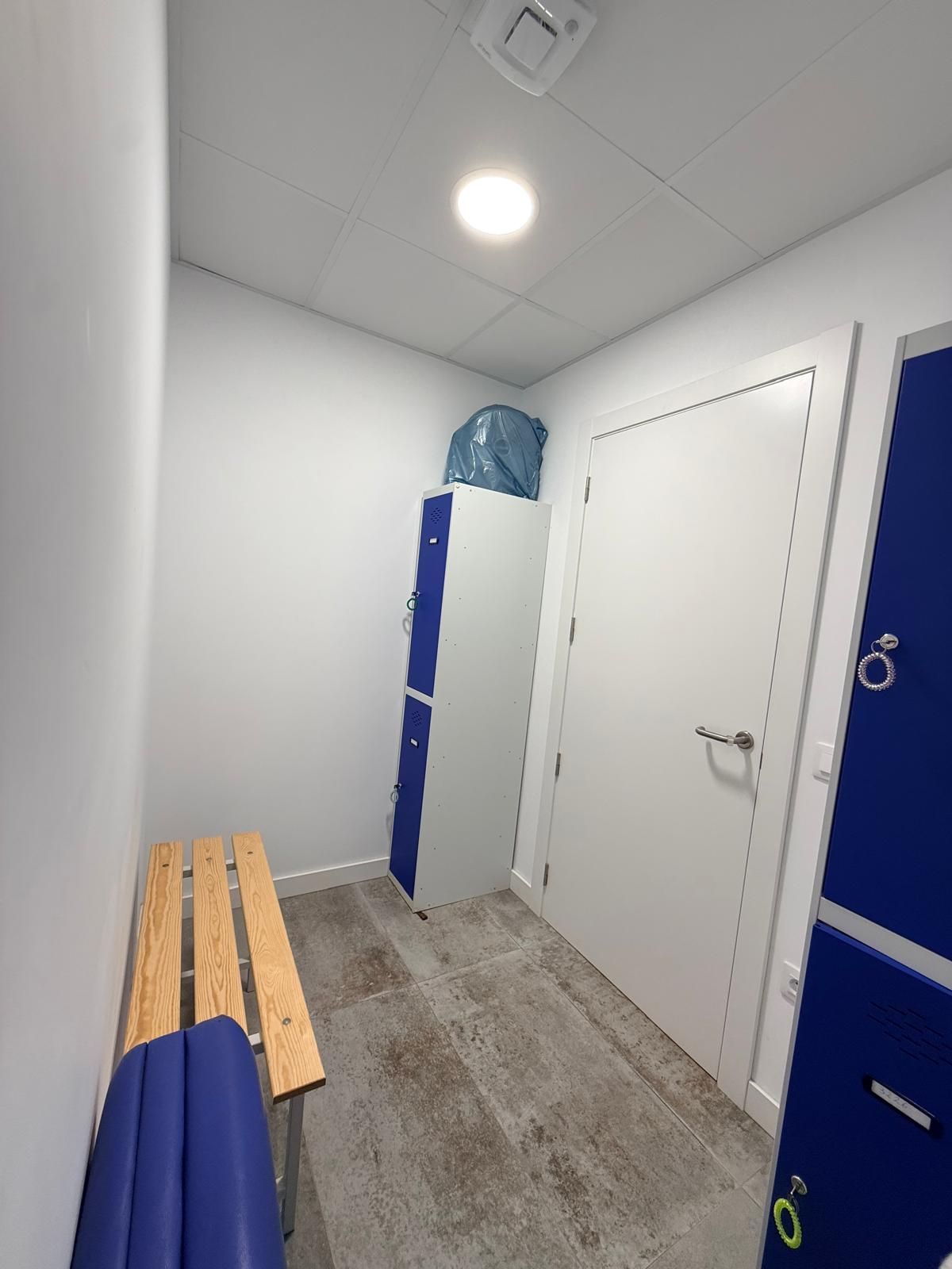 Vestuario estrecho con paredes blancas, puertas azules, un banco y suelo de baldosas bajo una brillante luz de techo.