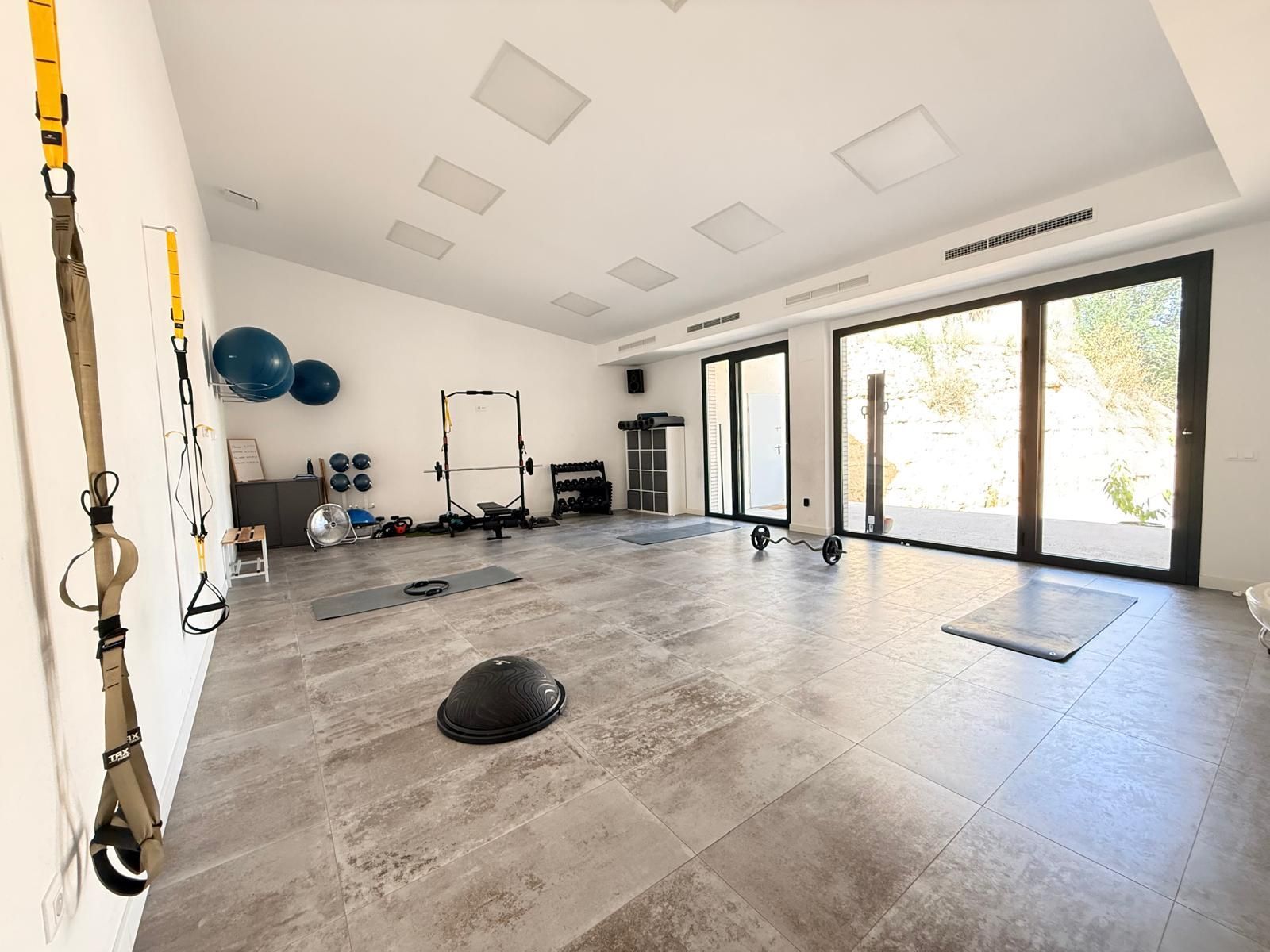 Gimnasio luminoso en casa con aparatos de ejercicio, colchoneta negra y grandes puertas de cristal que dan a un patio trasero.