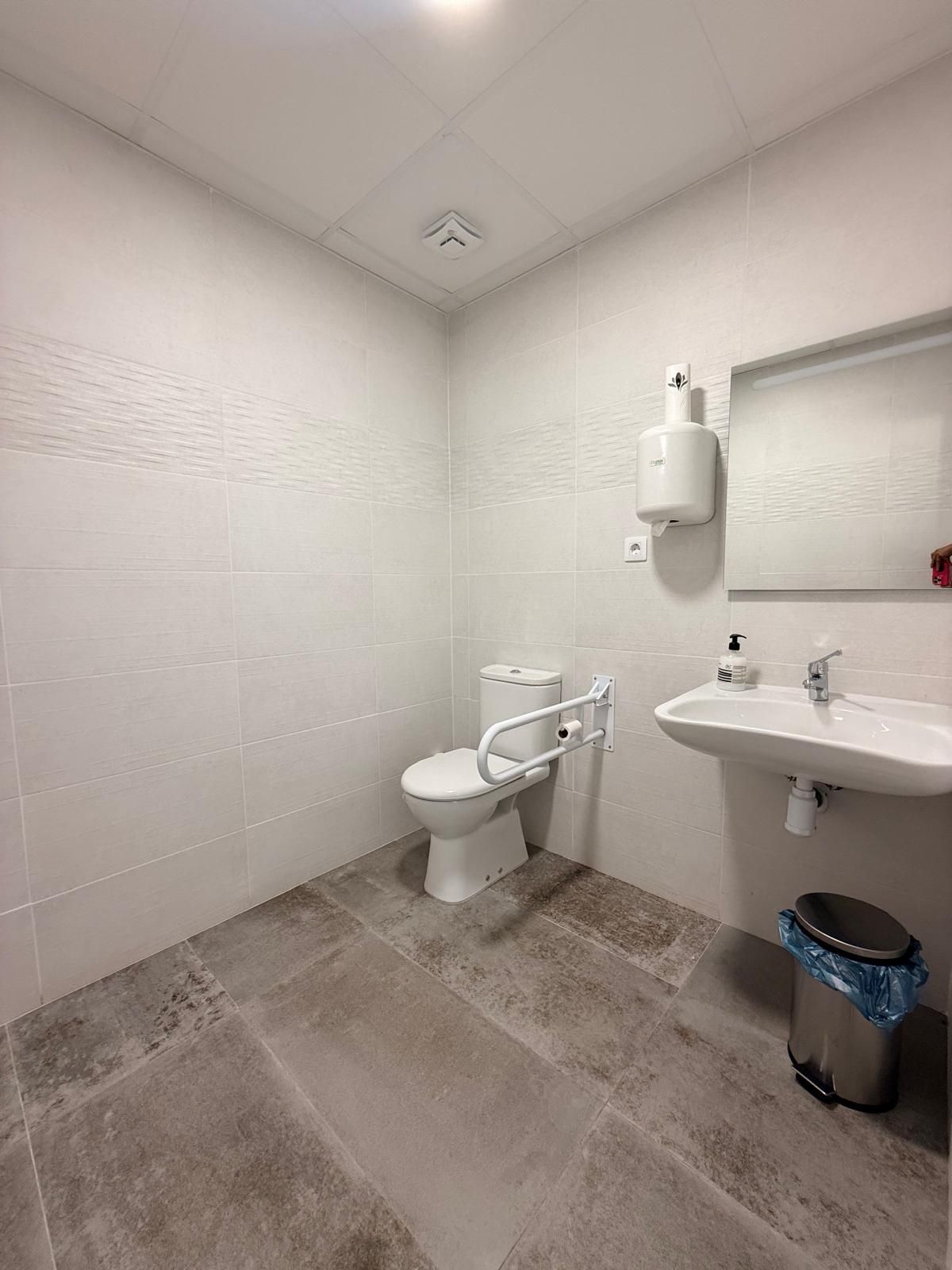 Baño accesible con paredes de azulejos blancos, inodoro, lavabo, barras de apoyo y papelera.