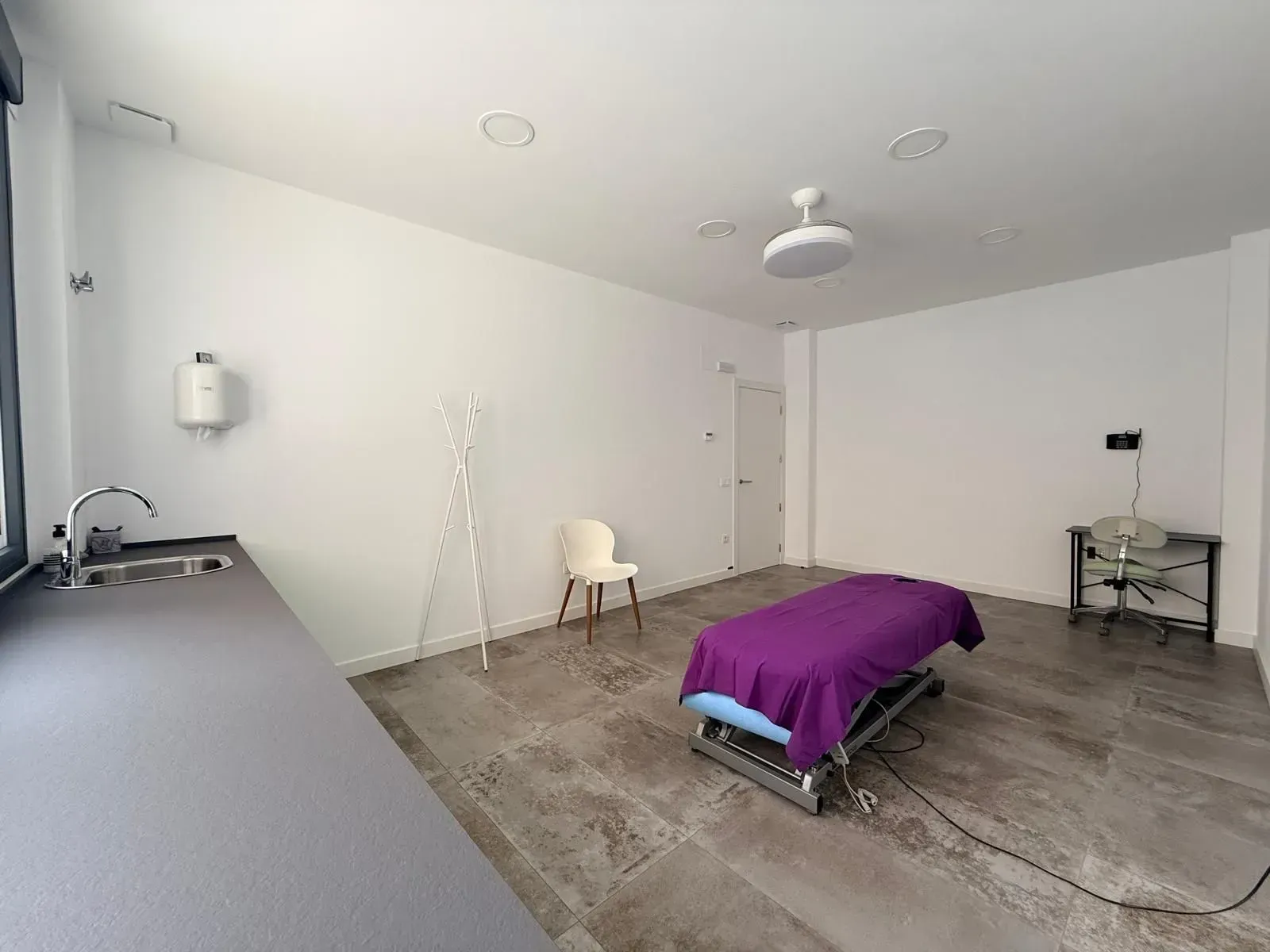 Una sala de tratamiento sencilla con una camilla de masaje, una silla, un lavabo y un pequeño escritorio en un espacio de paredes blancas.