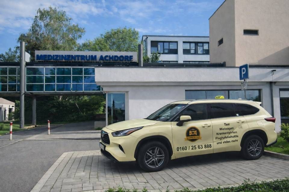Ein gelbes Taxi parkt vor dem Gebäude des „Medizinzentrums Achdorf“. Blaues Schild, moderne Architektur.