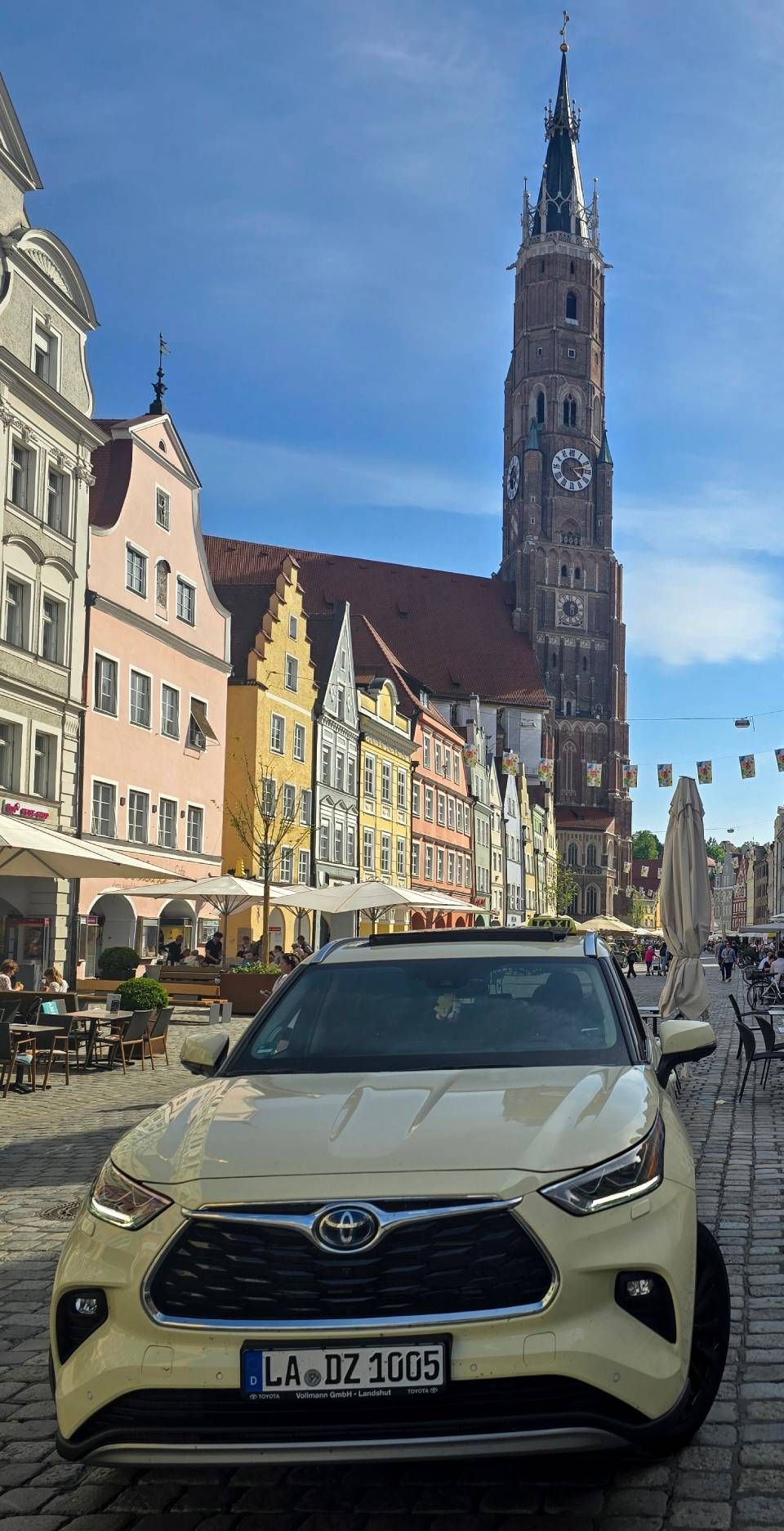Ein gelber Geländewagen parkt auf einer Kopfsteinpflasterstraße vor farbenfrohen Gebäuden und einem hohen Kirchturm.