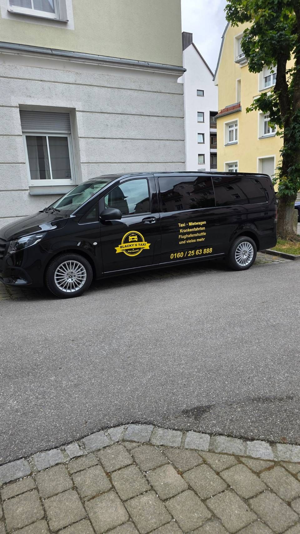 Schwarzer Lieferwagen mit gelber Aufschrift parkt auf einer gepflasterten Straße neben einem hellen Gebäude.