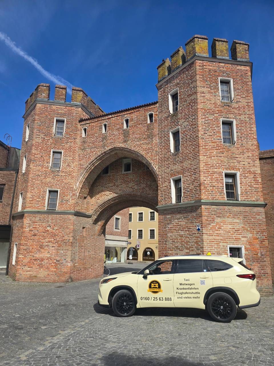 Ein Taxi parkt unter einem Backsteinbogen; zwei Burgtürme ragen zu beiden Seiten empor. Im Hintergrund blauer Himmel.