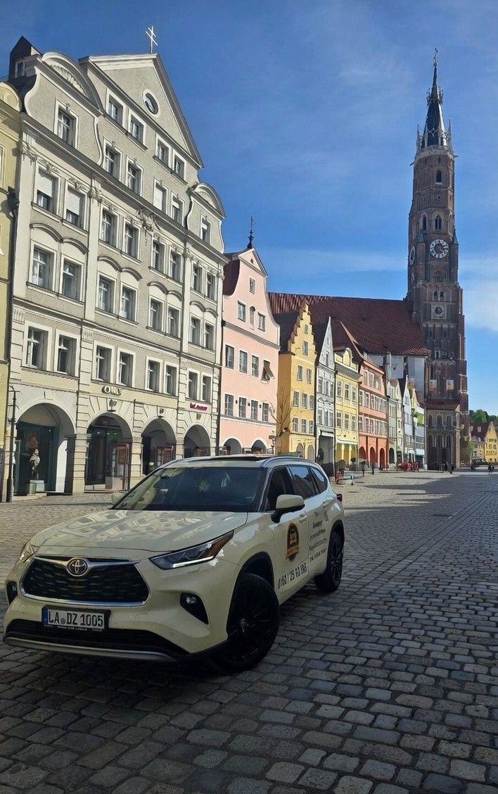 Ein Taxi parkt in einer Kopfsteinpflasterstraße vor farbenfrohen Gebäuden und einem Kirchturm. Sonniger Tag.