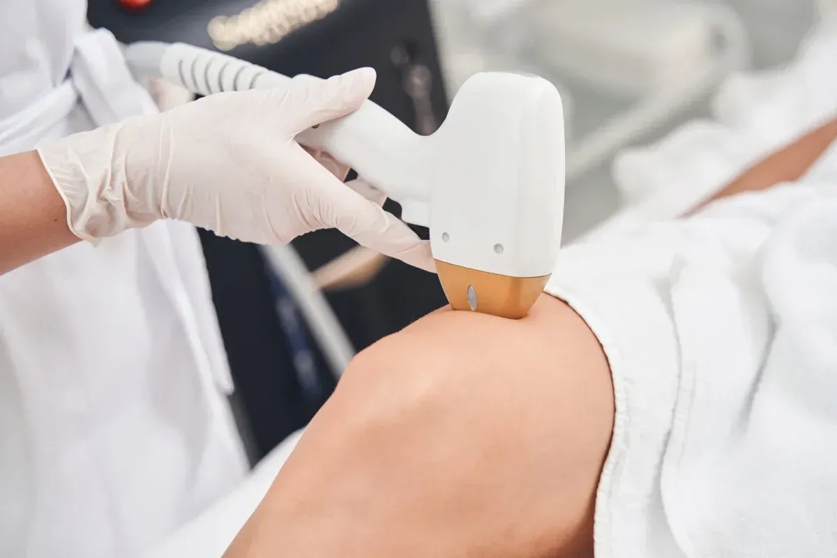 Un profesional, con guantes blancos, utiliza un dispositivo de depilación láser sobre la piel de una persona.