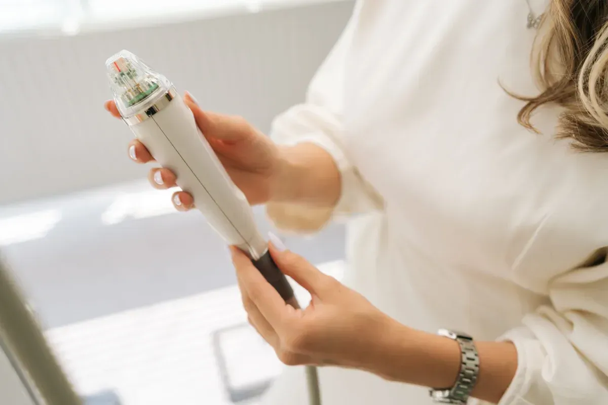 Una persona con una blusa blanca sostiene un dispositivo estético blanco de mano con una punta que emite luz.