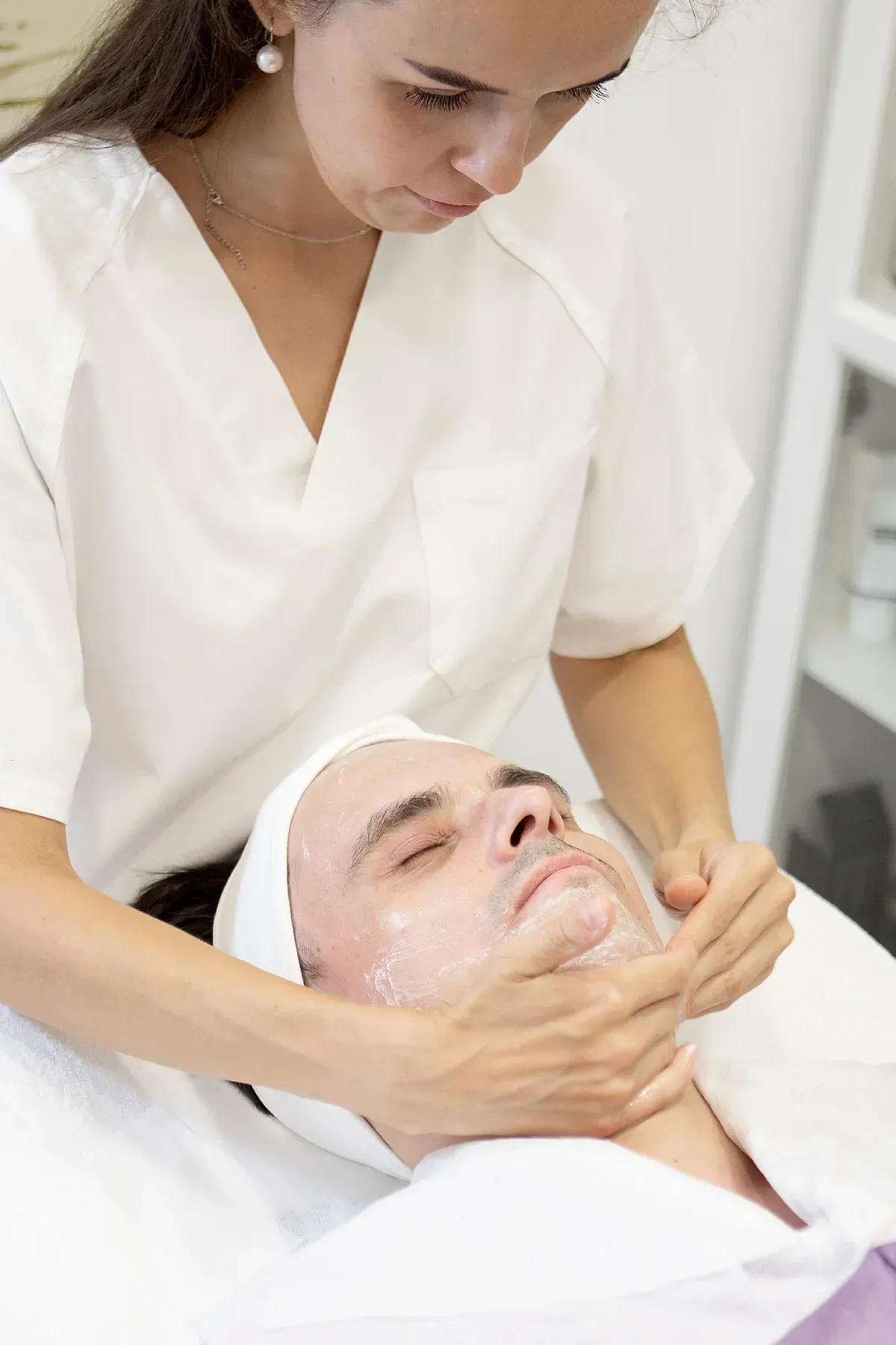Una persona con uniforme blanco realiza un tratamiento facial a una clienta que está tumbada en un spa.