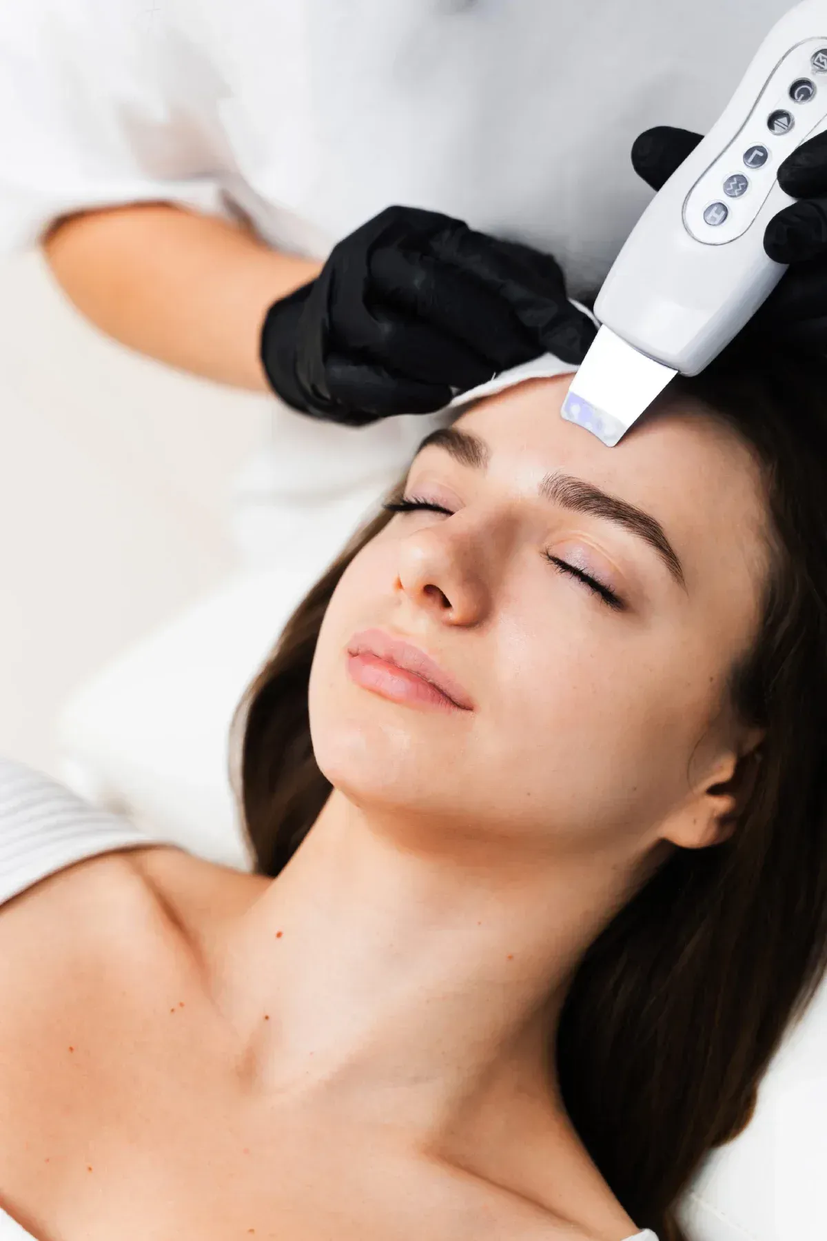 Una persona con una blusa blanca utiliza una espátula ultrasónica para exfoliar la frente de una clienta