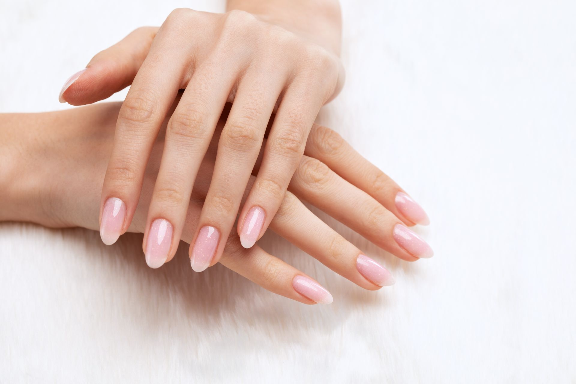 Deux mains avec des ongles ovales manucurés rose clair croisées sur une surface blanche et duveteuse.
