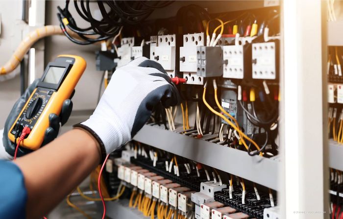 Elektriker mit Handschuhen testet mit einem Multimeter die Verkabelung der Schalttafel.