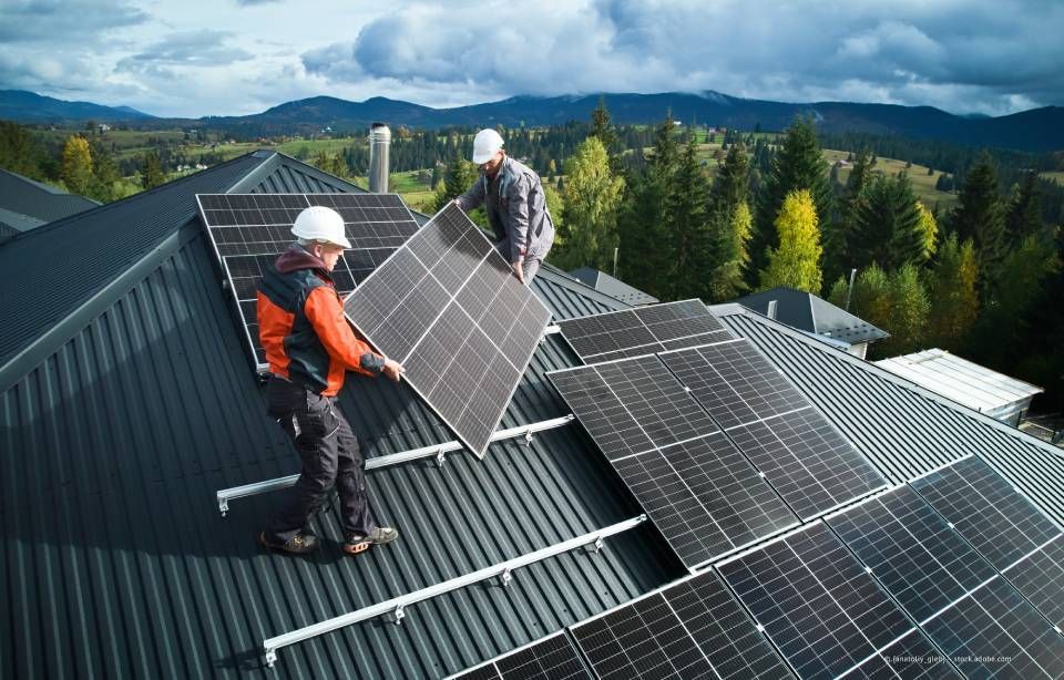 Zwei Arbeiter installieren Solarmodule auf einem Dach, umgeben von Bäumen, blauer Himmel.