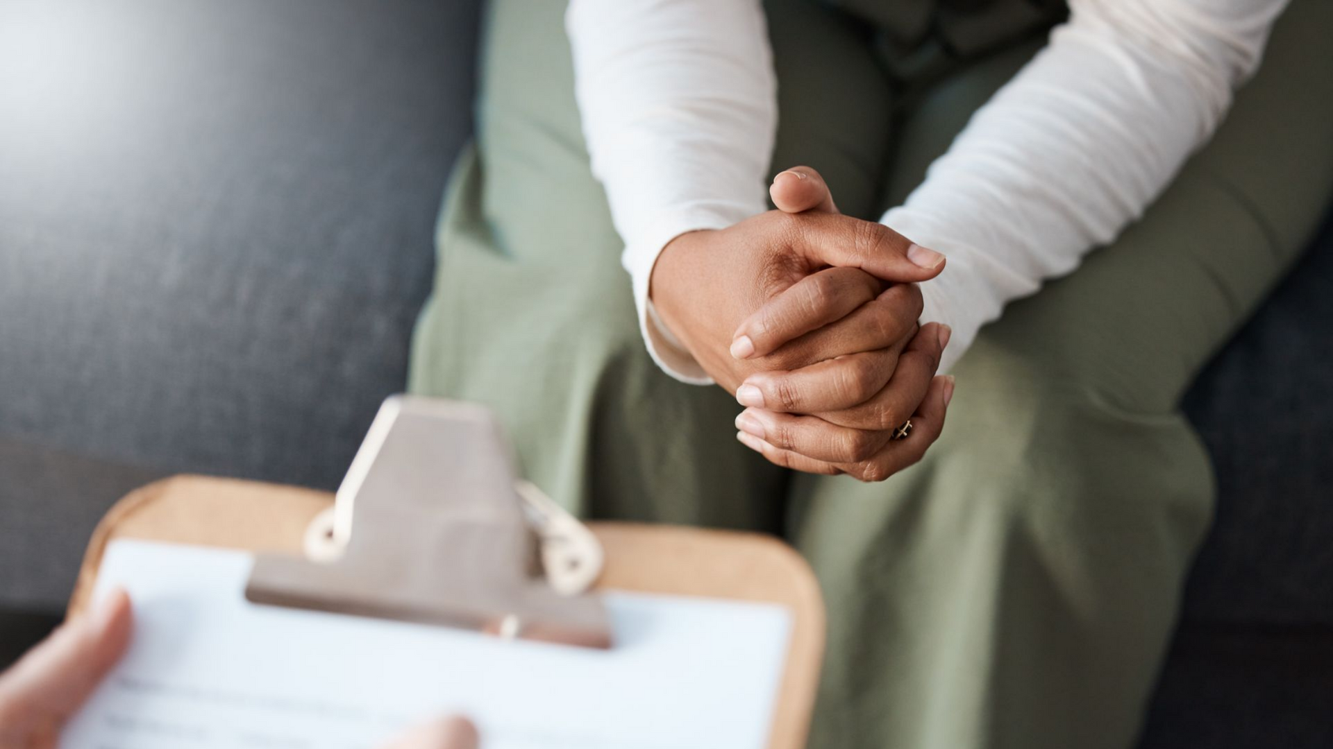 Mains jointes sur un bloc-notes ; personne en chemise blanche, pantalon vert, assise pour l’entretien.