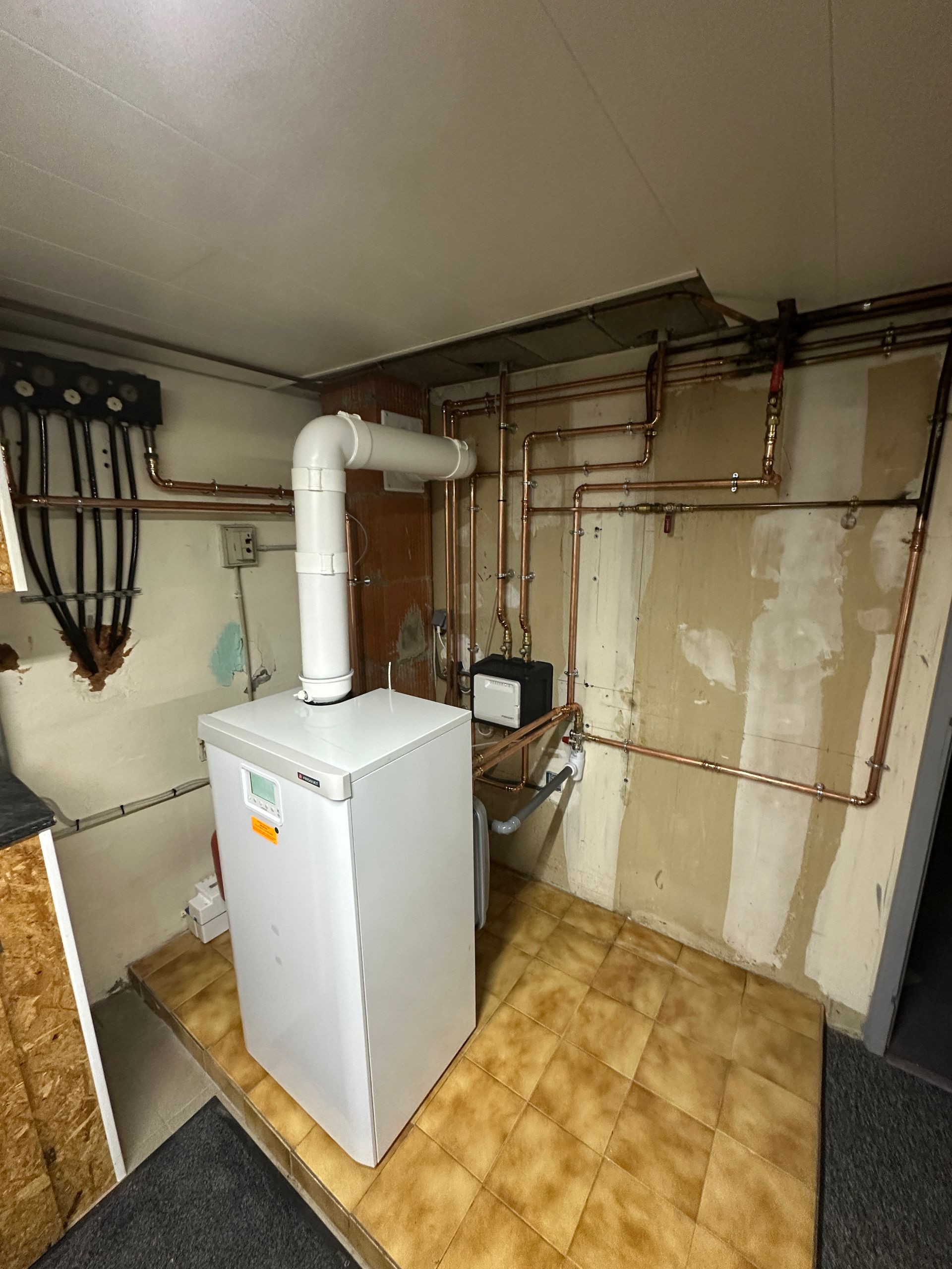 Chaudière blanche avec tuyau d'évacuation, tuyaux en cuivre et collecteur de distribution fixé au mur.