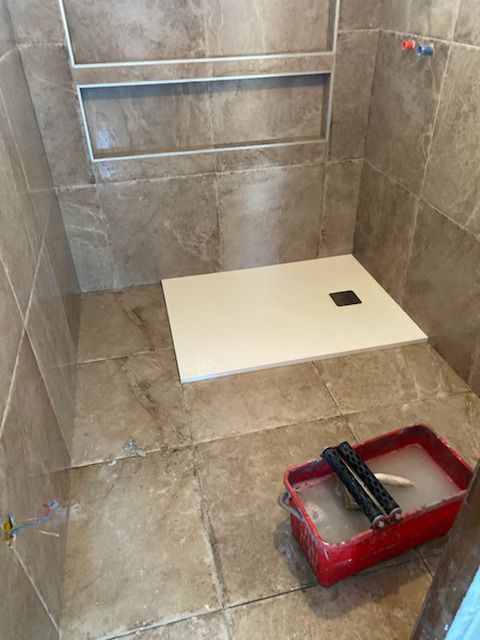 Cabine de douche en construction avec murs carrelés, sol blanc et seau rouge contenant des outils.
