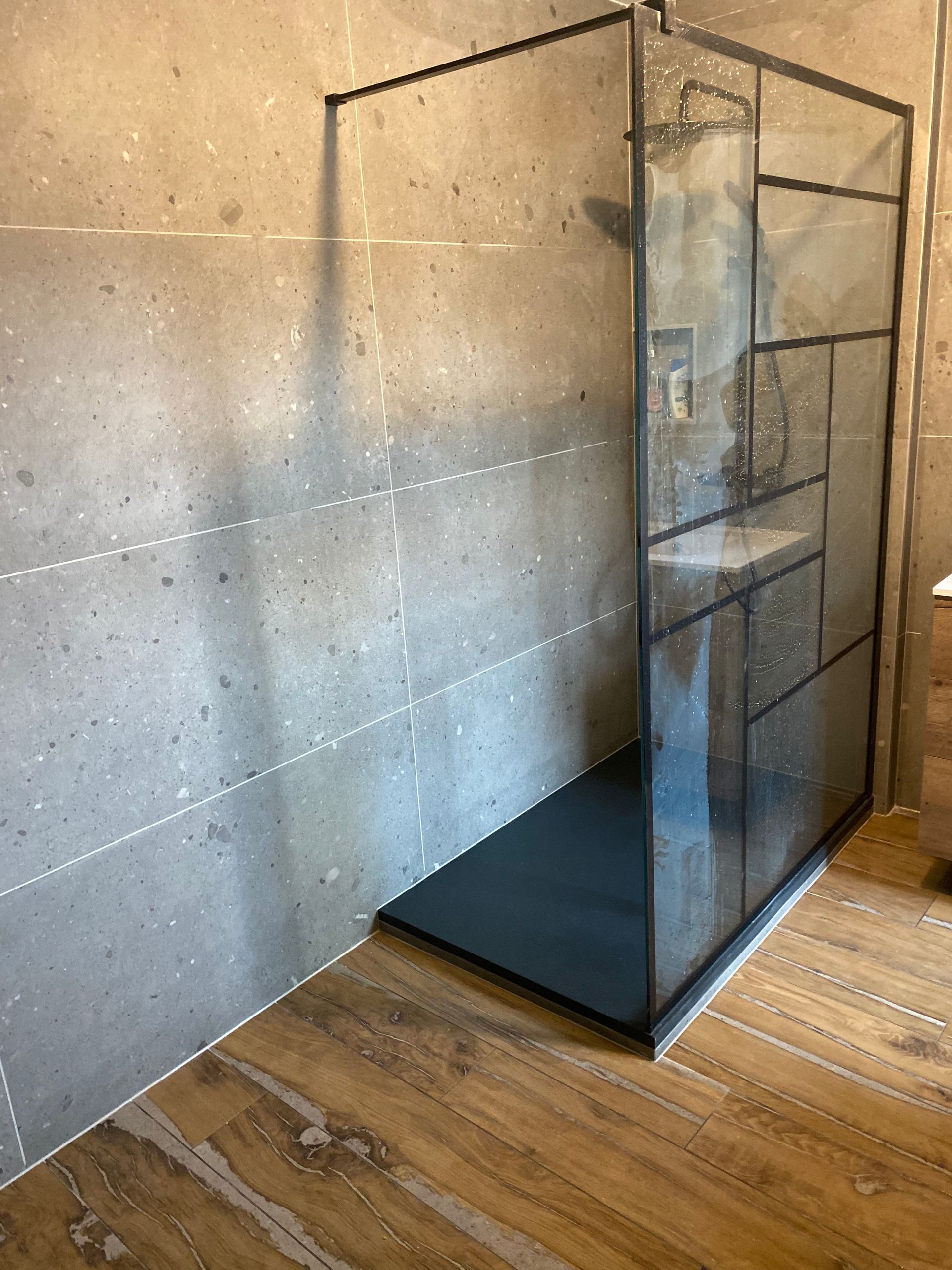 Une cabine de douche moderne avec un cadre foncé et des panneaux de verre, contrastant avec des murs carrelés gris et un sol en bois.
