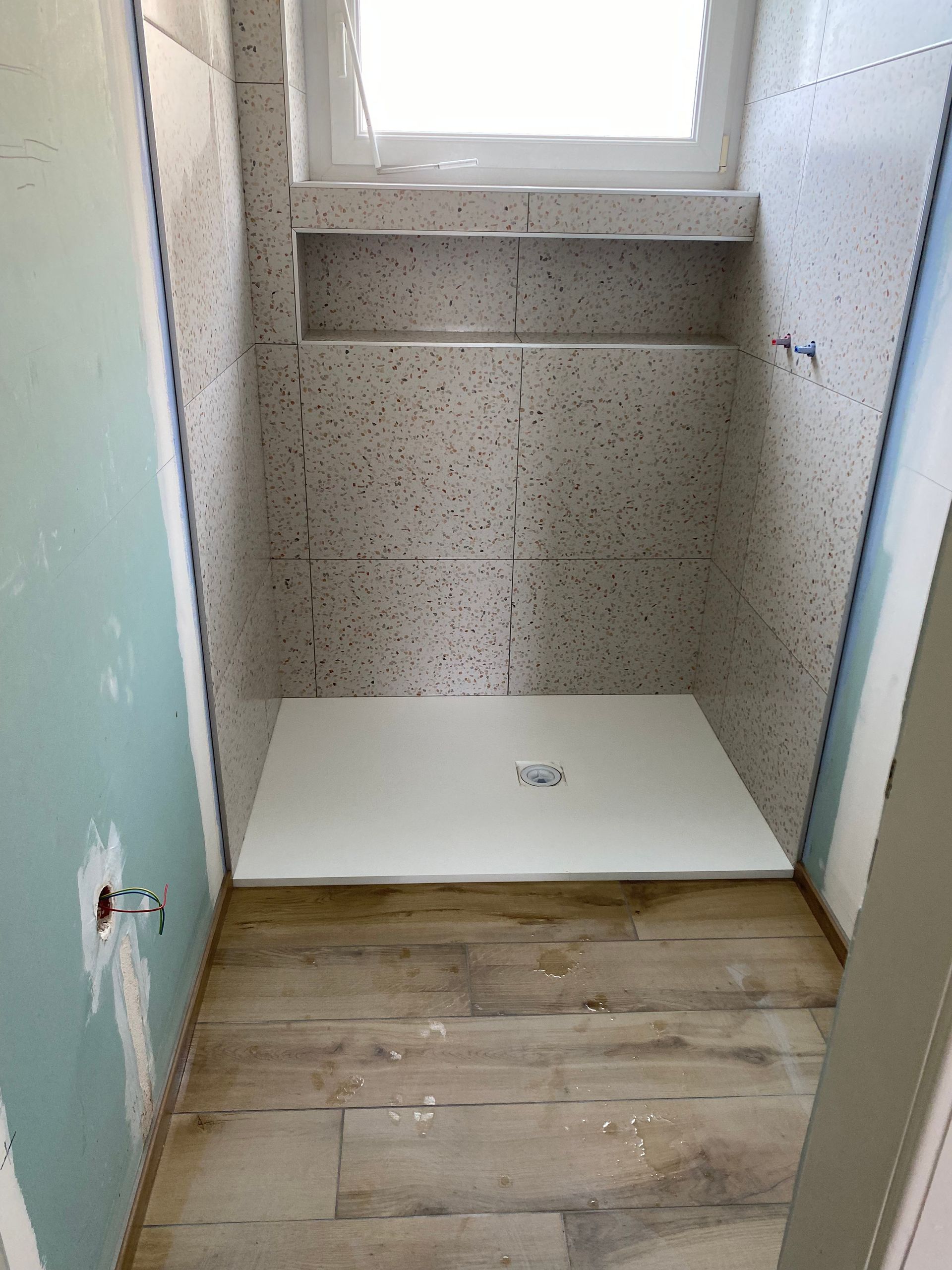 Rénovation de la salle de bain en cours : receveur de douche installé, murs carrelés, fenêtre au-dessus, sol imitation bois.