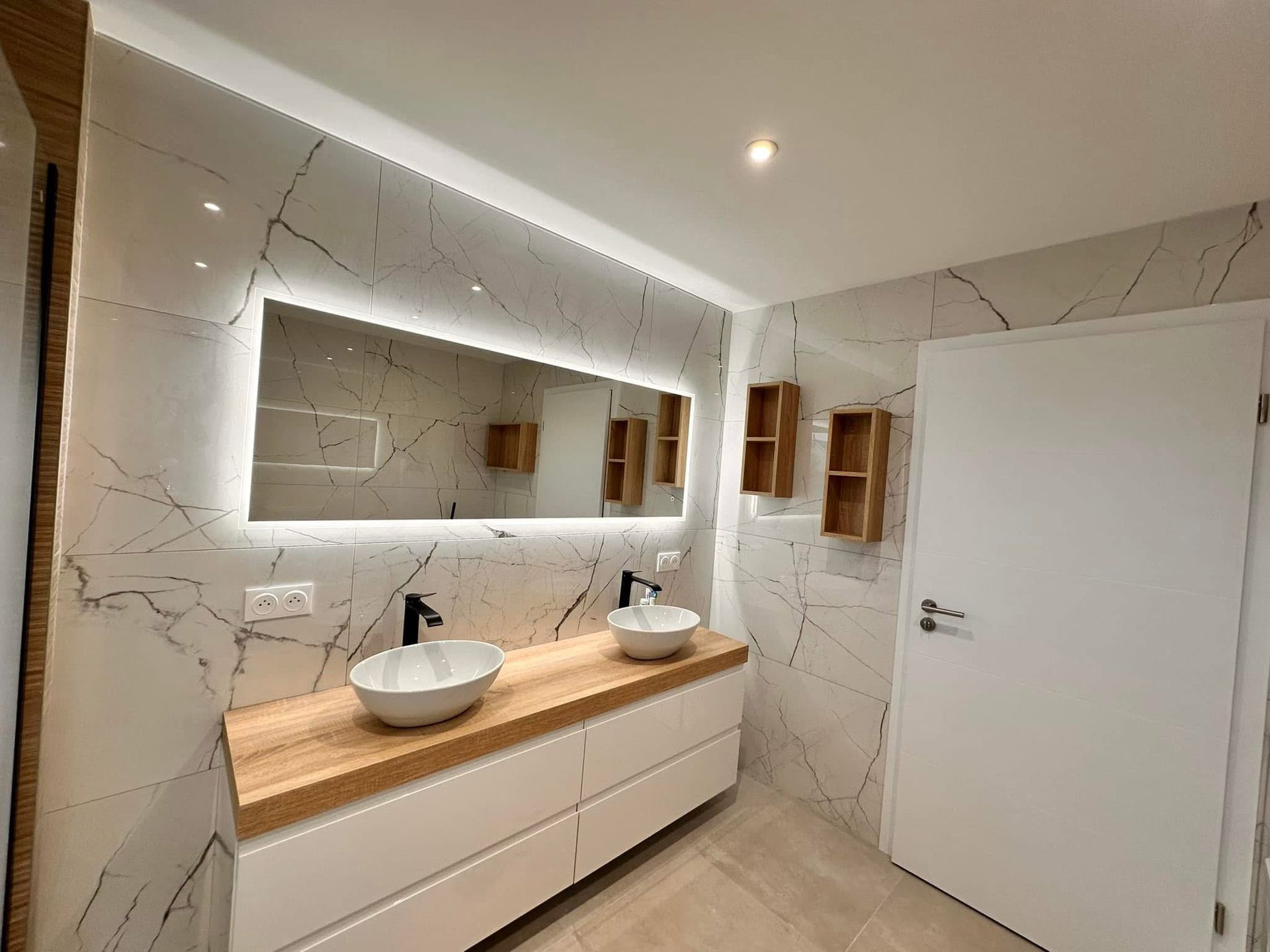 Salle de bains moderne avec murs imitation marbre, double vasque, grand miroir avec éclairage LED et éléments en bois.