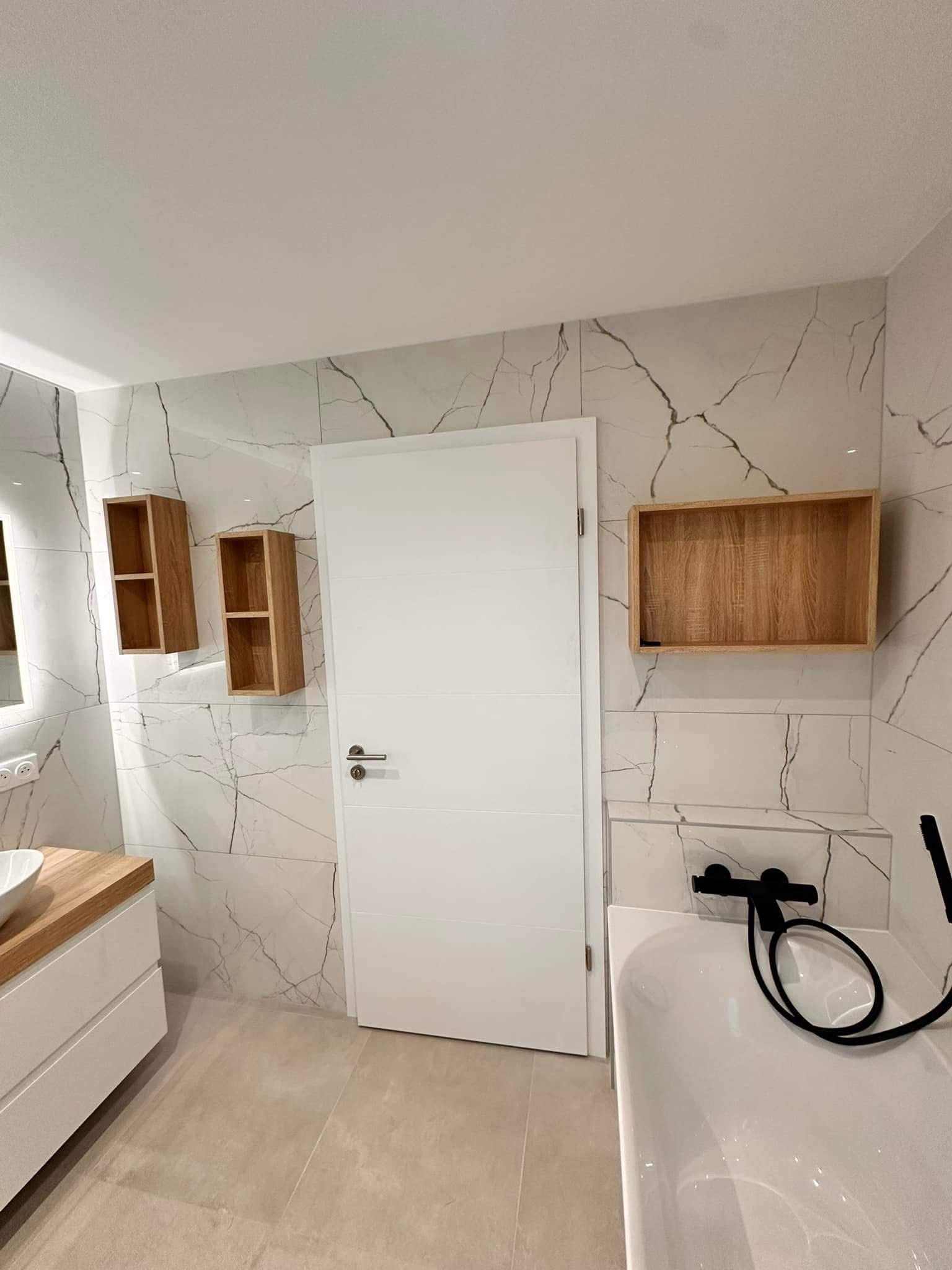 Salle de bains moderne aux tons blancs et boisés ; on aperçoit une porte, des étagères et une baignoire.