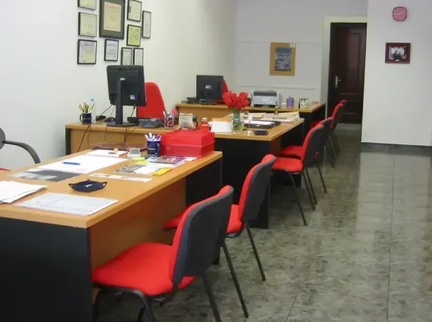 Oficina con escritorios, sillas rojas, computadoras e impresora. Certificados colgados en la pared.