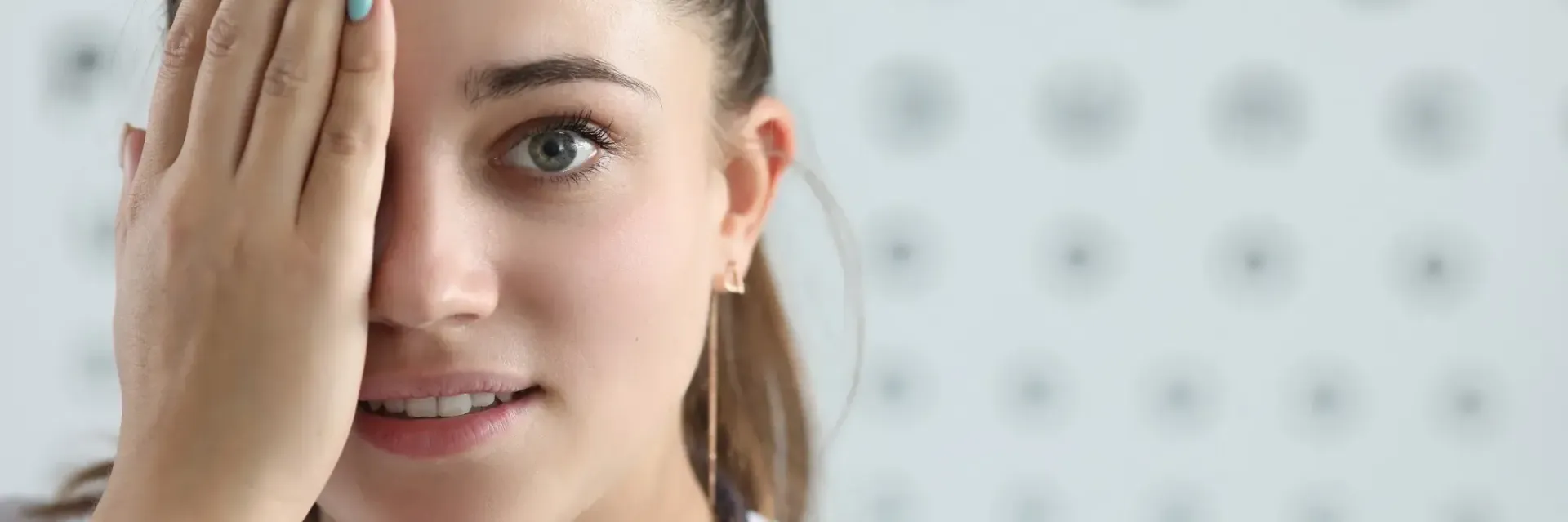 Una mujer se cubre un ojo durante un examen de la vista, sonriendo a la cámara. Al fondo, una pared gris claro.